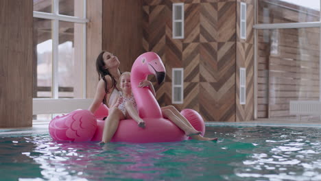 careful-mother-and-cute-little-girl-are-floating-on-inflatable-flamingo-in-swimming-pool-family-in-water-park
